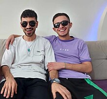 From left to right, (L-R) Matin Mannani and Hossein Naderbeigi sitting together on a white couch. Matin is wearing a white shirt and Hossein is in purple. Both are wearing sunglasses and laughing, light refracted on the wall behind them like a small rainbow.