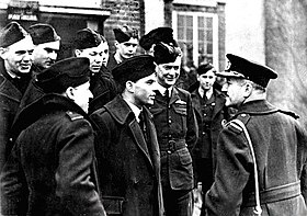 Half-length outdoor portrait of moustachioed man in military great coat with peaked cap, talking to a group of ten or men in military uniforms with forage caps