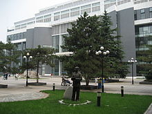 large bâtiment de verre et béton, 6 étages. esplanade avec lampadaires et quelques résineux. statue en bronze d'un homme en col mao.