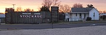 Log stockade with low square towers at corners, bearing large sign "Medicine Lodge Stockade", next to one-story frame house with wraparound porch