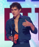A long shot of a young man with curly brown hair, wearing a blue unbuttoned shirt. He is accepting an award on stage and speaking into a microphone, looking into the audience.