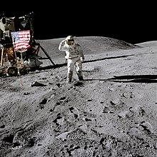 Photographie en couleur de Charles Duke à côté le drapeau américain. Le rover lunaire et le module lunaire sont visibles derrière le drapeau.