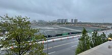 An empty, level, gravel construction site on a rainy day. The site is surrounded by fencing, with ad boards showing renders of the proposed stadium project.