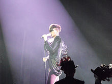Photograph of a woman who sings on stage. She wears a dark leather blouse and a short skirt, and light tights.