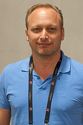 A close-up picture of a smiling man facing the camera in a blue polo shirt wearing a lanyard around his neck.