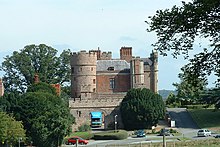 A large brick building built in the style of a medieval castle; grass, bushes and some trees in the foreground.