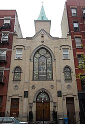 The front of a modest 3-story beige church, between 2 brick apartment buildings, seen from across the street, looking upward. A steeple is visible toward the back.