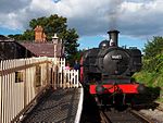 A black pannier tank locomotive is standing at the platform of a small rural railway station with a brick and slate building, cream wooden fencing, and lamp. The driver is standing next to the locomotive and is talking to a group of passengers who are about to board the red passenger carriage.