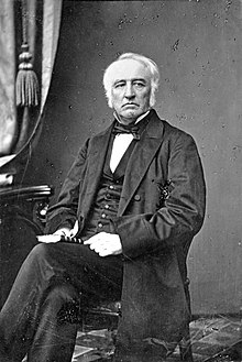 Fair-skinned, white-haired man, sitting in a formal pose, wearing mid-Victorian suit
