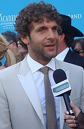 A man with dark curly hair and noticeable beard stubble, wearing a light grey jacket and dark grey tie, talking into a microphone