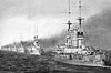A long line of large, light gray warships sail through calm waters, each belching thick black smoke