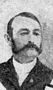 Portrait of a white man with a combover and sideburns connected to a mustache, wearing a dark suit.