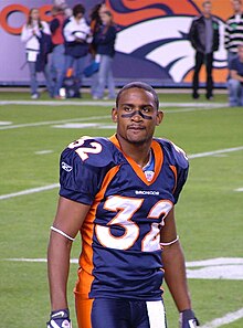 An American football player looks at the crowd. He is wearing his uniform on the field with some spectators in the background.