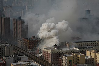 An elevated view of the fire following the explosion, with a plume of smoke, fire department trucks with ladders extended, and firefighters hosing water from the ladder buckets and from the street. The elevated railroad is in front of the scene.