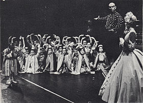 Black and white photo of a theatrical scene: a middle-aged man with a shaved head and imposing presence has his right arm extended to introduce a group of children in Asian dress to a woman in a crinoline dress and bonnet in the foreground at right, who is partially turned upstage. The children are mostly kneeling and have their arms raised in greeting; one child (probably Crown Prince Chulalongkorn) stands and bows.