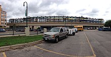 An elevated train station crosses a busy road in the background; in the foreground, a silver SUV is parked.