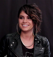 Still image of a woman looking away from the camera and smiling with an undercut hairstyle; wearing large hoop earrings, a black top, a thick necklace and a leather jacket.