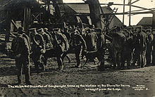 A uniformed stretcher party carry a body away from the pithead; others wait around the pithead. Wreckage from the explosion can be seen in the background.