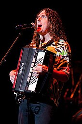 A photograph of "Weird Al" Yankovic, singing through a microphone and playing a harmonica.