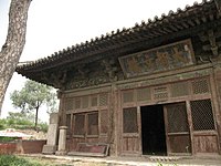 Half a of a red-walled building with large eaves projecting from the corner. A tree trunk is visible on the left-hand side of the image