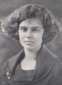 A young white woman with thick wavy bobbed hair, wearing a dark dress with a cowl neckline