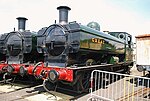 Two pannier tank locomotives are seen from the front and to the right and are standing next to each other in front of a turntable. Both locomotives have black chimneys and fronts. The cabs, pannier tanks, clackbox covers and boiler domes are all green. The buffer beams are red and include the locomotive number in yellow with black edging, 7752 on the left locomotive and 7760 on the right. On the right locomotive, one round cab window is visible and is edged in brass.