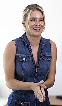 A smiling blonde woman in a sleeveless jean jacket and a visible cochlear implant with palms pressed together at a right angle to her body.