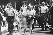 Black-and-white photograph of two women with shaved heads and blank expressions on their face walking down a street in Paris. The women are surrounded by a group of other people, most of whom are smiling.