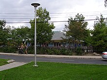 one-story wooden building with outdoor seating and parking lot. Trees surround the seating area and a small road is situated nearby.