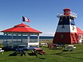 Photographie du centre d'information touristique de Grande-Anse. Le bâtiment, en forme de phare, est orné du drapeau de l'Acadie. Il y a près de celui-ci une gloriette et des tables de pique-nique peints aux couleurs du drapeau de l'Acadie. Un drapeau de l'Acadie est également monté au sommet de la gloriette.