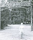 Woman in a long dress in front of a sign across a road. Wooden letters read "Camp Curry".