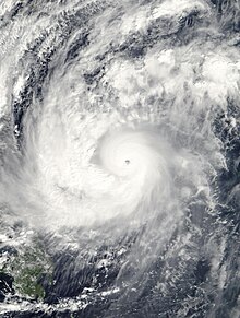 A well organized tropical cyclone, centered at bottom right, with a well-defined eye and spiraling rainbands. A landmass is visible under heavy cloud cover at left.