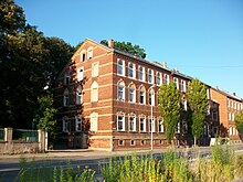 zwei viersöckige Häuser, dreispurige Straße (davon zwei Spuren mit Pfeilmarkierungen zum Abbiegen), Grünbewuchs