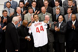 Several rows of men standing and clapping; in the front is a smiling, brown-skinned man holding a white baseball jersey with "Obama" and a large "44" in red on the rear toward camera