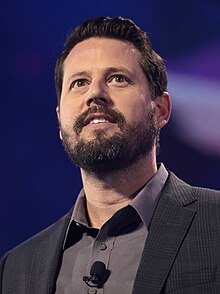 Portrait photograph of Seth Dillon from the shoulders up, wearing a white shirt and blue jacket, holding a microphone