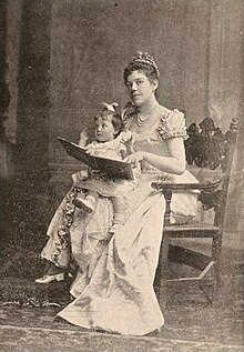 Woman wearing a ruffled gown and tiarra, seated on a chair, and holding a child and a book