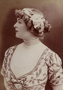 photographie en noir et blanc d'une femme en buste, au port de tête altier, ayant la chevelure ornée de rubans, vue de profil gauche