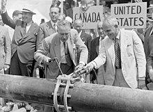 Two men are attaching a piece of metal to a pipeline. A crowd is gathered behind them