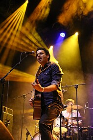 A curly-haired man wearing a blue shirt sings into a microphone strums and strums an acoustic guitar