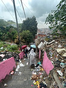 Pessoas descendo uma escadaria com uma grande quantidade de lixo e entulho nos matos nos dois lados. As pessoas estão de costas e com guarda-chuvas.