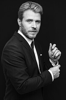 black-and-white image of white male from profile, wearing dark suit, buttoning a cuff, and looking at camera