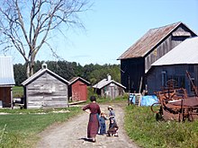 An Amish woman and three children, on a path to a house and six wooden farm buildings, past some farm equipment