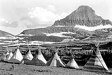 A black-and-white photograph. Against a backdrop of mountains and forests is a row of eight tipis.