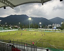 A grass football pitch with a stand on one side. Next to it other pitches are visible. There are mountains in the background and dramatic clouds in the sky.