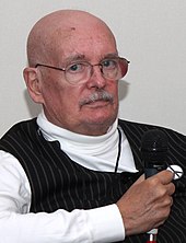 Dennis O'Neil, a bald man wearing sunglasses, a plaid blue shirt, and a gray vest, smiles at the camera.
