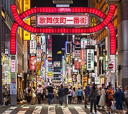 Red lighted gate denoting entrance to Kabukichō, a district in Shinjuku-ku, Tokyo. Colorful neon signs for businesses line both sides of the street.