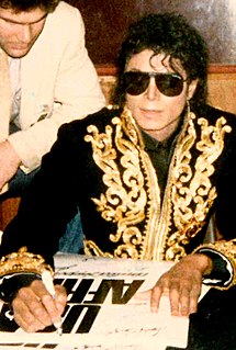 Jackson is sitting at a table autographing a 'We Are The World'poster. He's wearing sun glasses and a black jacket with a golden appliqué on the front and the cuffs. The zip is half open. Jackson is wearing a dark shirt under the jacket He's looking straight into the camera. His skin is light brown.