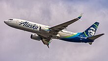 A plane painted with the words 'Alaska' in the front and a blue-green eskimo on the tail soars after take-off just under a cloudy sky