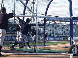 A man in gray baseball pants and a navy blue top swinging a bat at a baseball in a batting cage