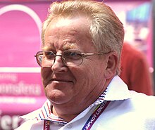 A middle-aged short-haired blond man with strong complexion is wearing a white polo shirt under a red vest; his eyes are closed and he has glasses hanging from his collar. Other people face stand behind him facing towards the background with some tents and trees.